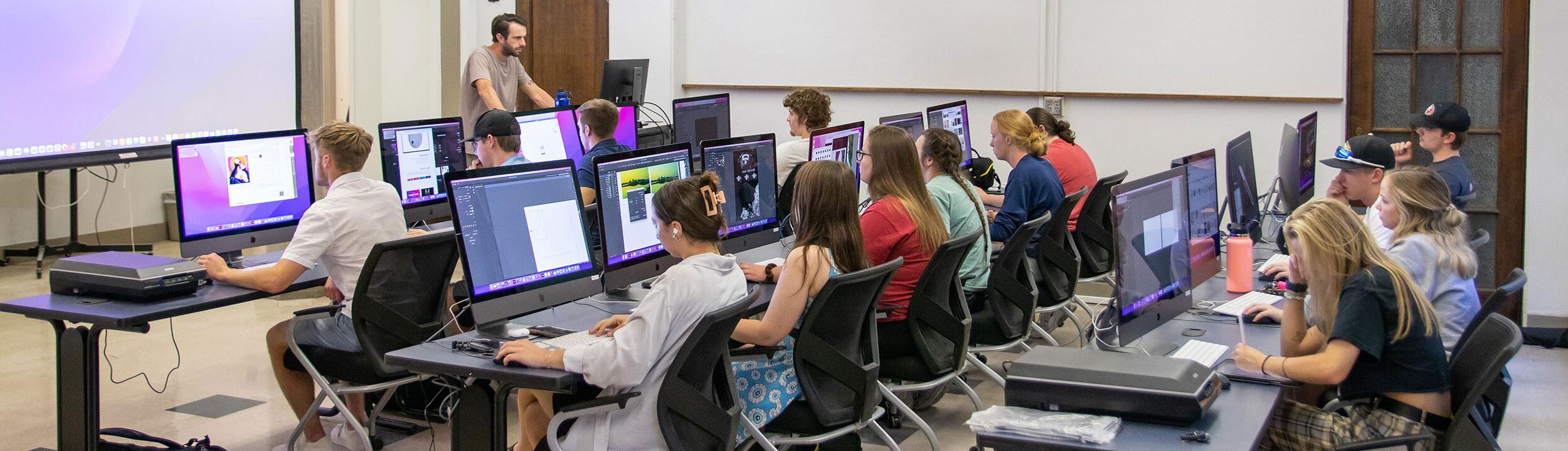 教室里有三排桌子和大显示器的电脑. 学生们坐在课桌前，老师站在教室前面.